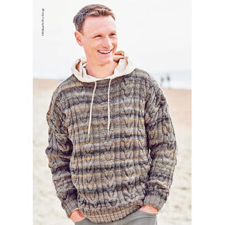Mann in gemustertem Pullover und Kapuze sitzt am Strand mit Lächeln.