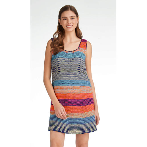 Frau in buntem, gestreiftem Long-Top steht lächelnd vor einem hellen Hintergrund.