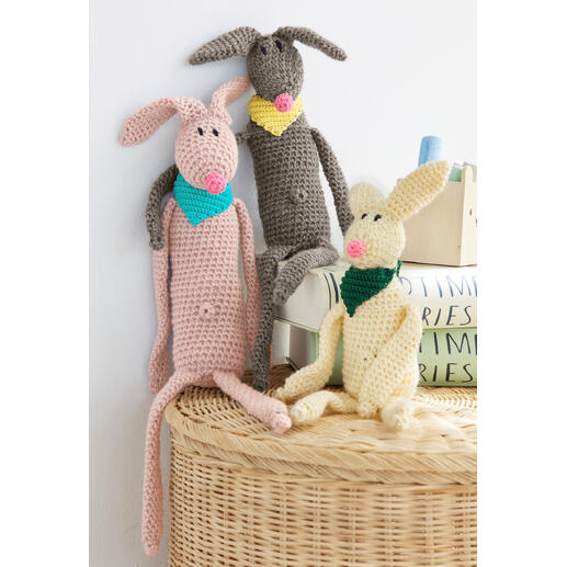 Drei gehäkelte Hasen in verschiedenen Farben, mit Halstüchern, auf einer Korboberfläche.