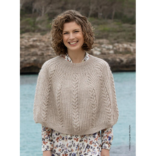 Lächelnde Frau in einem beigen Poncho aus dickem Garn vor einer Wasserfläche.