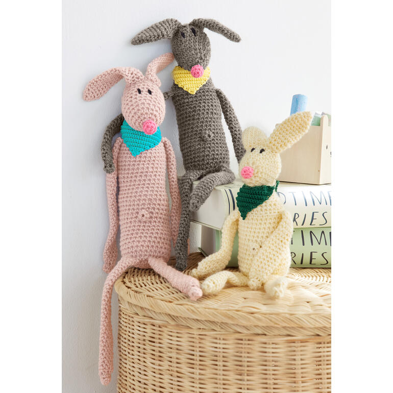 Drei gehäkelte Hasen in verschiedenen Farben, mit Halstüchern, auf einer Korboberfläche.