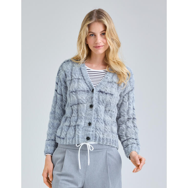 Frau in grauer, strukturierter Jacke mit Knöpfen, steht lächelnd vor einem neutralen Hintergrund.