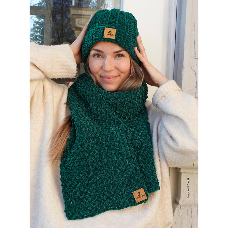Frau mit Schal und Mütze in Dunkelgrün, aus Samt gehäkelt, lächelt in die Kamera.