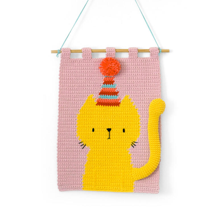 Häkel-Wandbehang mit gelber Katze und Partyhütchen auf rosa Hintergrund.