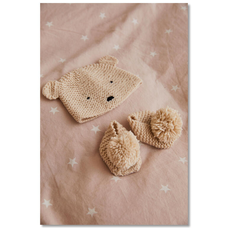 Bär-Mütze und -Schühchen aus Wolle auf einem rosa Stoff.