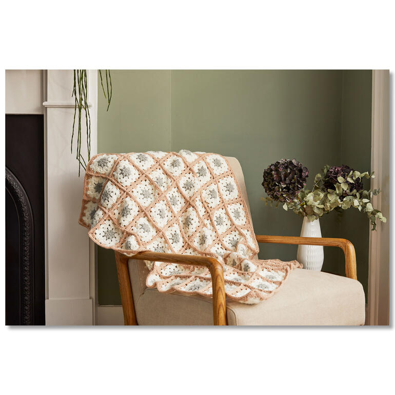Granny Square Decke auf einem Sessel drapiert neben einer Vase mit Blumen.