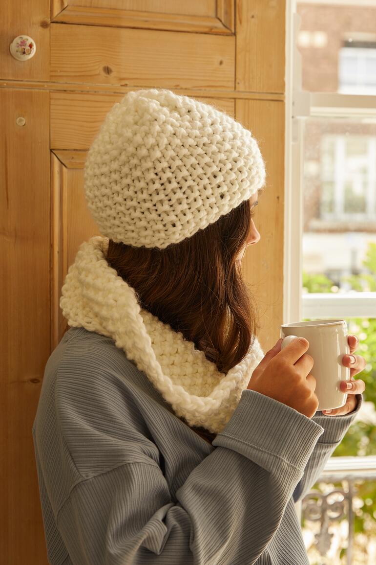 Frau mit einer weißen Mütze und Loop, hält eine Tasse.