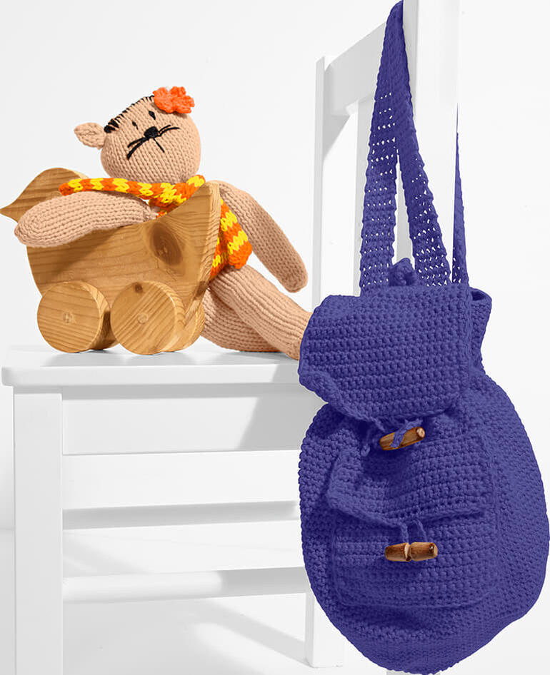 Gehäkelte Schlenkerkatze in Beige mit Blumenkopf auf einem Holzkorb, neben einem blauen Rucksack.