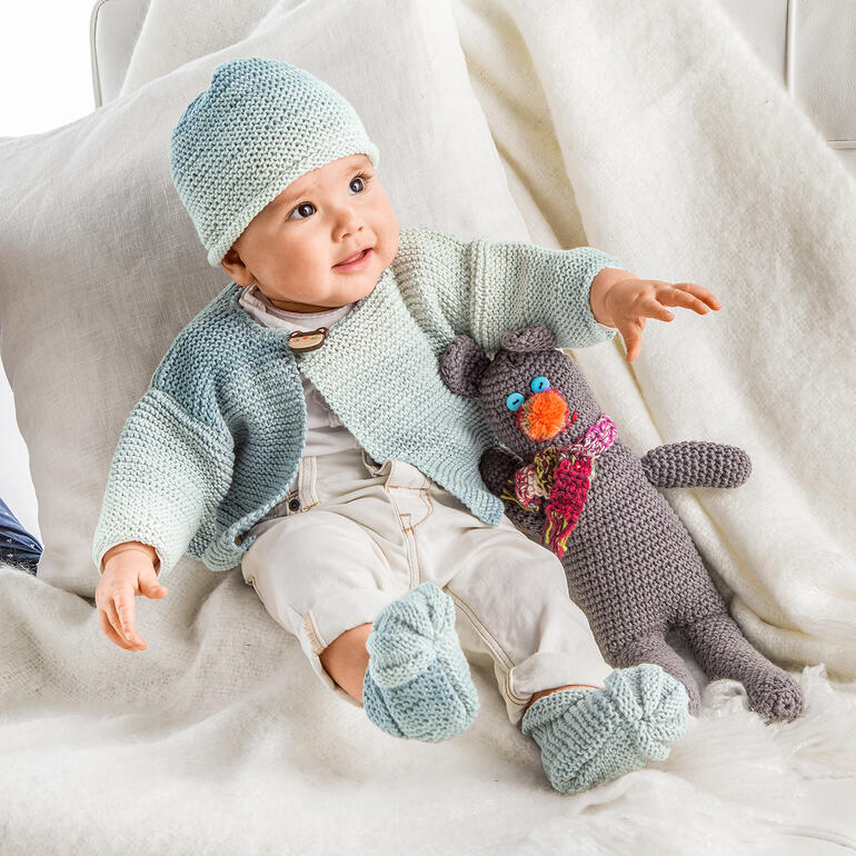 Baby-Jacke, Mütze und Schühchen in sanften Blautönen, zusammen mit einem Stofftier.