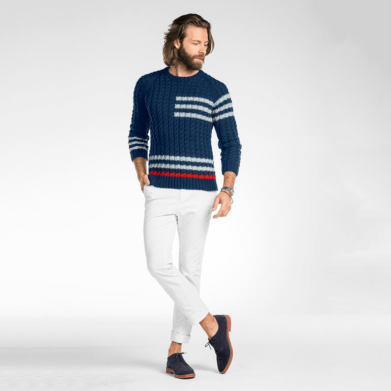 Mann trägt blauen Pullover aus Merino-Cotton mit Zopfmuster und weißen Hosen, steht auf einer hellen Fläche.