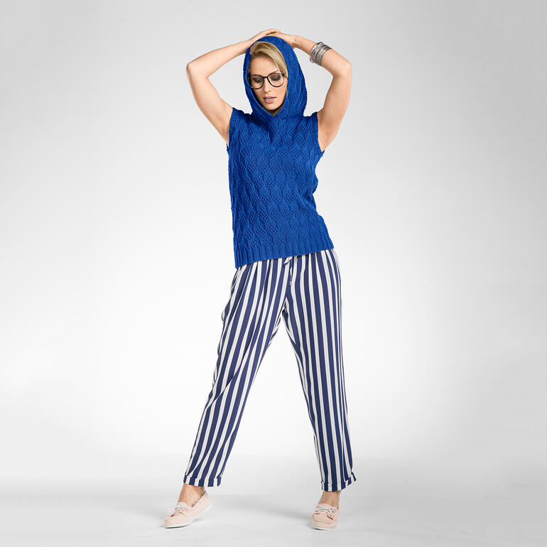 Frau in blauer ärmelloser Kapuzenpullover mit Zopfmuster und gestreiften Hosen, posiert mit erhobenem Arm.