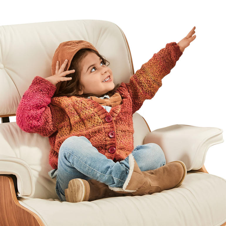 Kind in bunter Strickjacke, sitzt lächelnd auf einem Sessel.