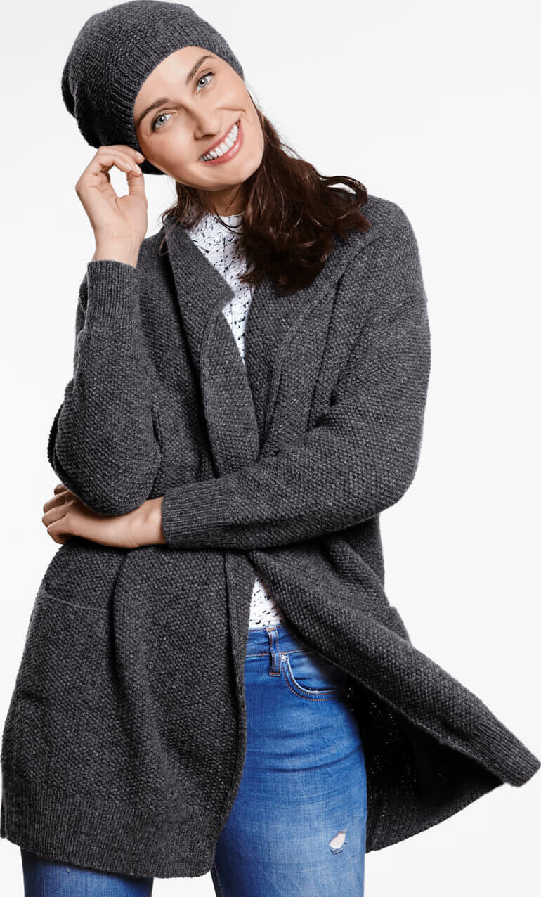 Dame in grauem Mantel und passender Mütze, lächelnd vor neutralem Hintergrund.