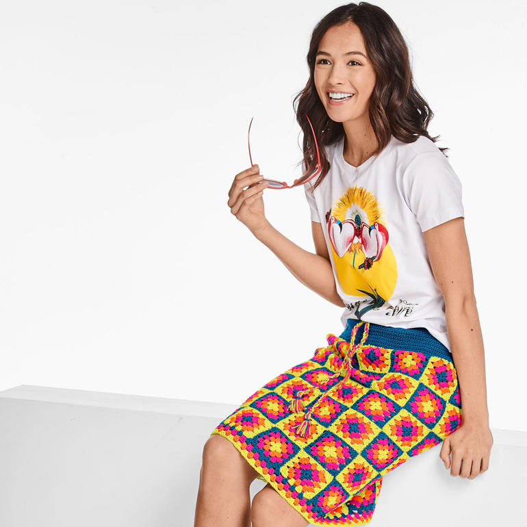 Lächelnde Frau in einem bunten Häkelrock mit einem T-Shirt und Sonnenbrille.