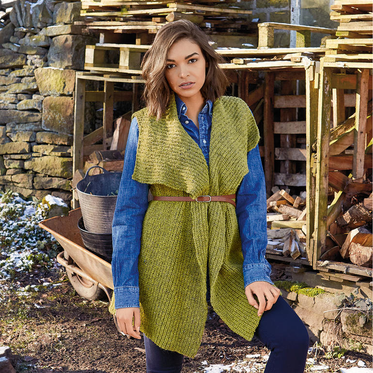 Frau in einer grünen Weste über einem blauen Hemd, mit einem Gürtel, vor einer Holzstruktur.