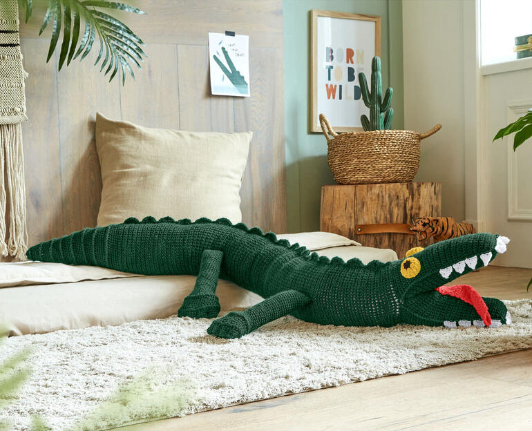Grünes gehäkeltes Krokodil, liegend auf einem Teppich im Raum.