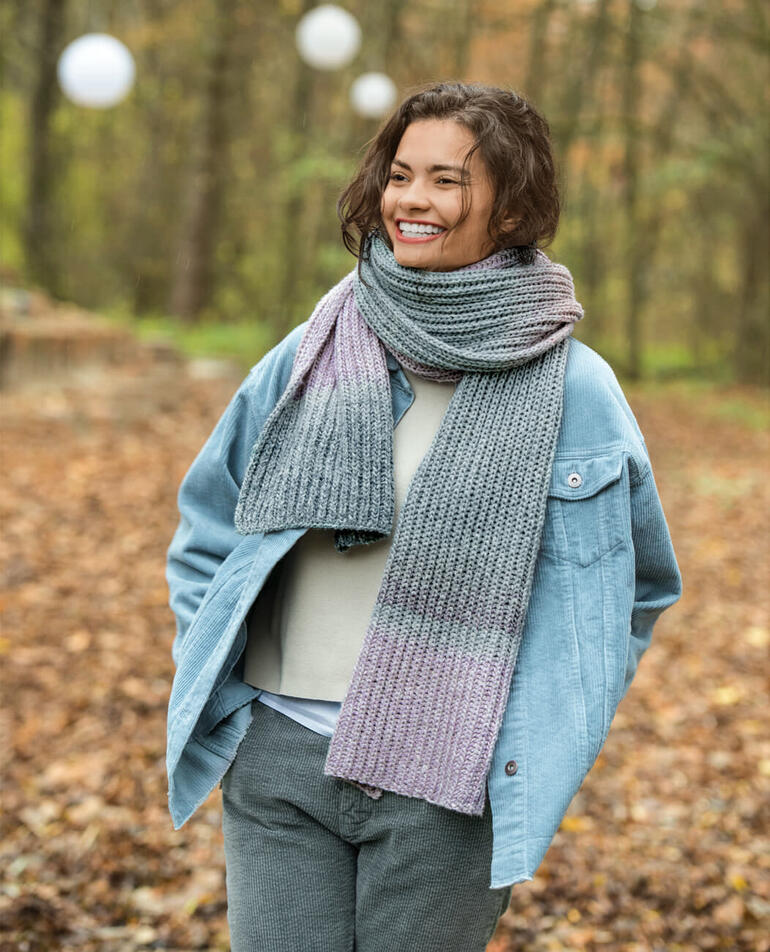 Frau trägt einen Schal aus Pastello in Grautönen, lächelt vor herbstlicher Kulisse.