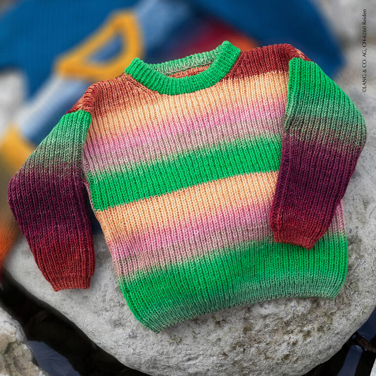 Bunter Merino-Pullover auf einem Stein, im Hintergrund weitere Pullover.