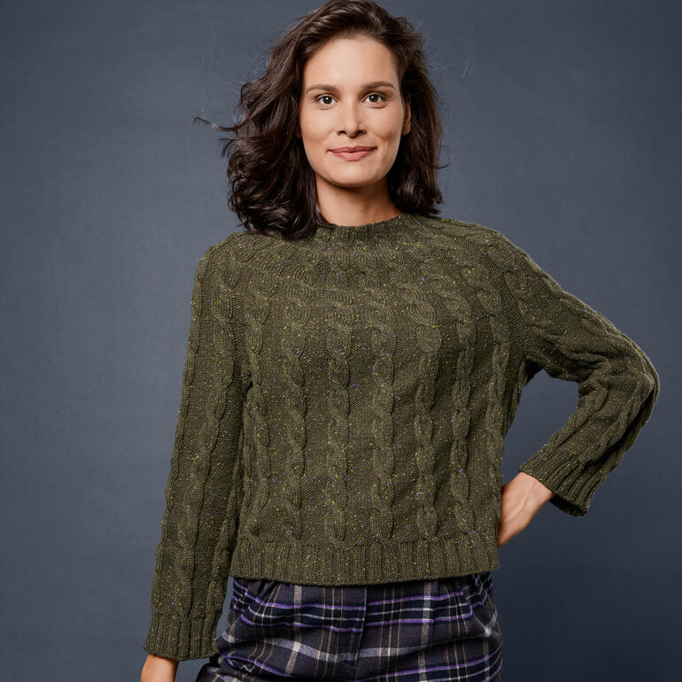 Frau trägt einen Cropped-Pullover aus Como Tweed, posiert vor grau-blauer Wand.