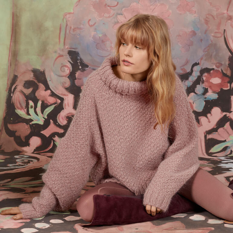 Frau in rosafarbenem Pullover aus Honor steht vor einem bunten Hintergrund.