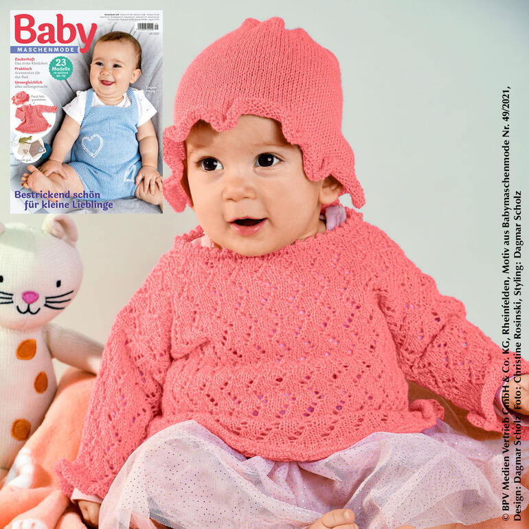 Baby trägt einen rosa Pulli und eine passende Mütze aus Seidana, sitzt lächelnd.