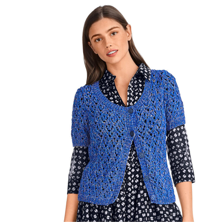 Frau in blauer gehäkelter Jacke aus Oletta, die während des Lächelns die Haare berührt.