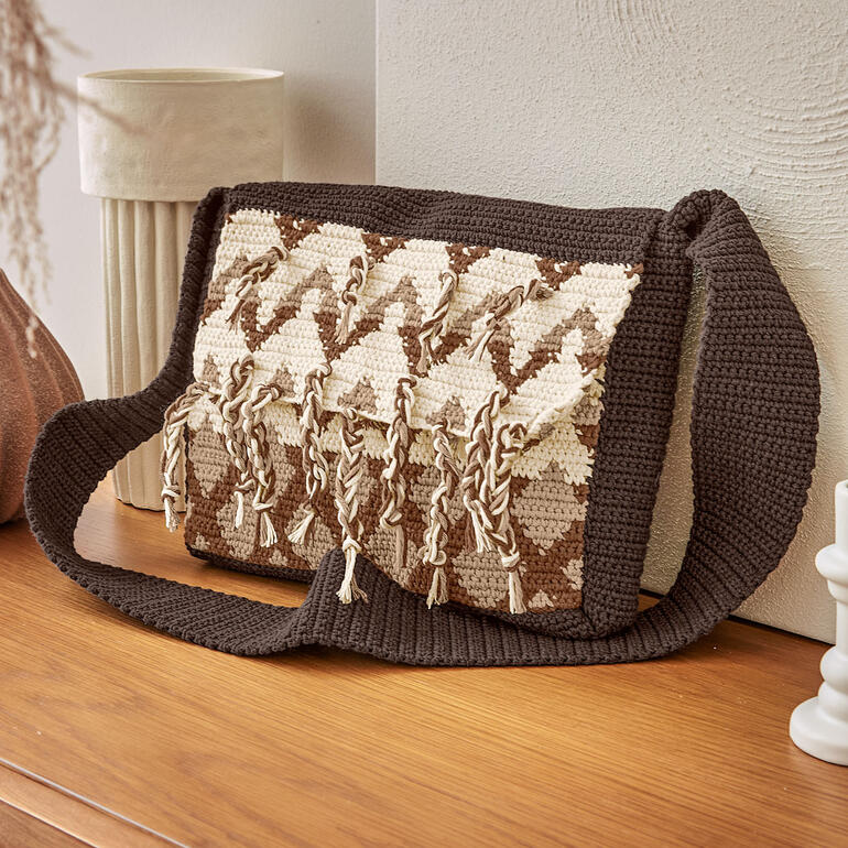 Braune gehäkelte Tasche mit Fransen und einem gestreiften Muster auf einem Holztisch.