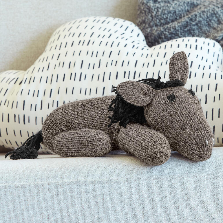 Gestricktes Kuscheltier Pferdchen liegt auf einem hellen Sofa mit Kissen im Hintergrund.