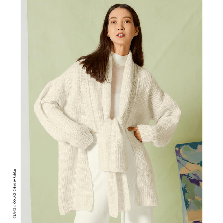 Frau in cremefarbener Jacke mit Gürtel vor einer grünen Wand.