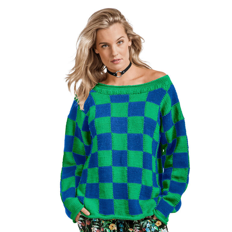 Frau in grün-blauem Pullover aus Cotonara steht auf einer weißen Fläche.