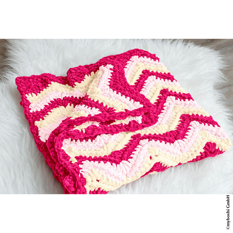 Häkeldecke in pink und cremefarbenen Wellenmustern auf einem weißen Untergrund.
