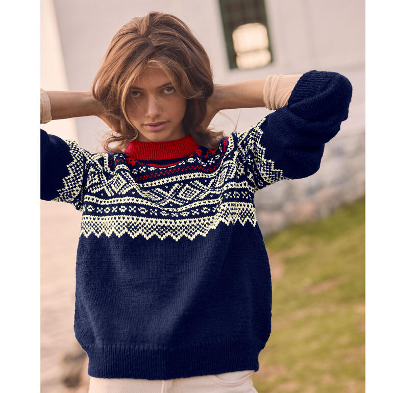 Frau in blauem Pullover mit nordischem Muster vor grünem Hintergrund, spielt mit Haaren.