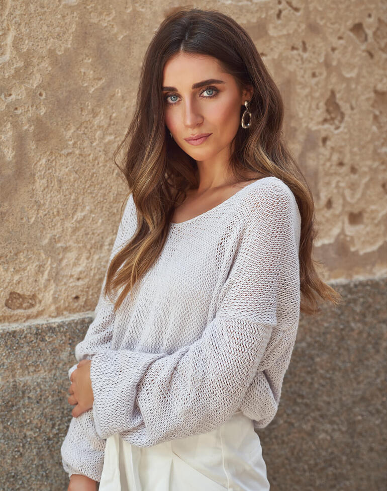 Frau in hellgrauem Pullover mit weitem Ausschnitt steht vor einer Wand.