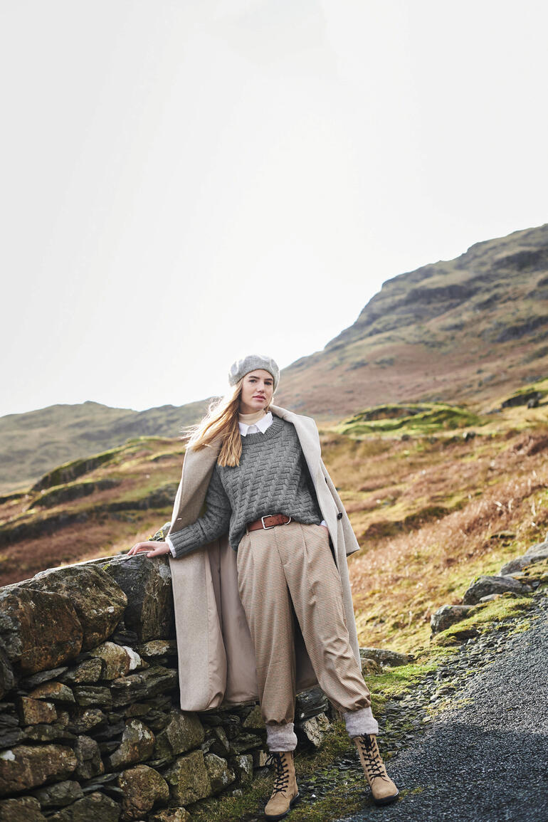 Frau in grauem Pullover und beige Camel Coat vor hellem Hintergrund