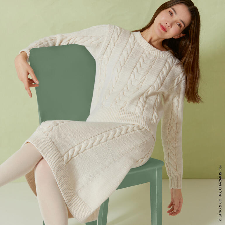 Pullover aus Merino+ in cremefarben, getragen von einer Frau auf einem Stuhl.