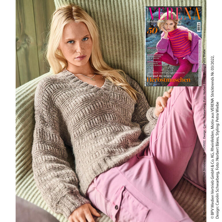 Blonde Frau trägt einen beigen Pullover auf einem Sofa, mit einer Zeitschrift in der Hand.