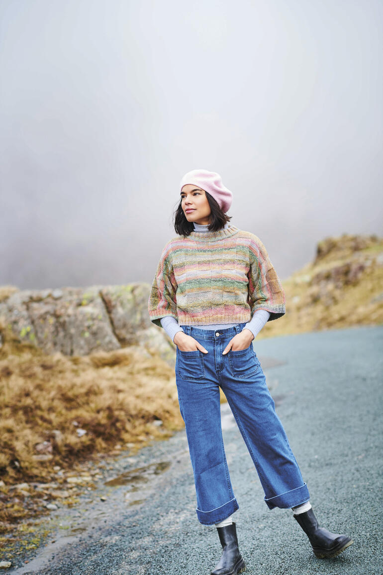 Frau in gestreiftem Pullover und rosa Mütze posiert in der Natur.