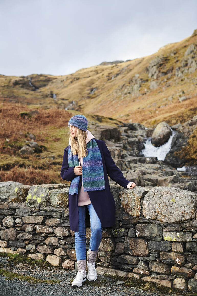 Blonde Frau trägt eine Mütze und einen Schal in Blau- und Grüntönen im Freien.