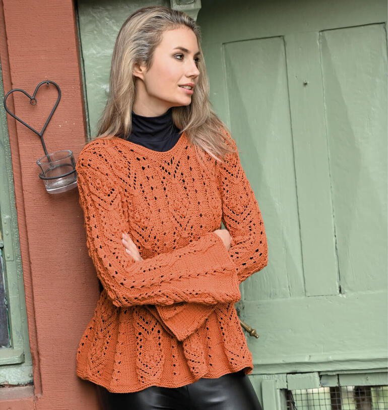 Frau in orangefarbenem Strickpullover lehnt lässig an einer Wand.
