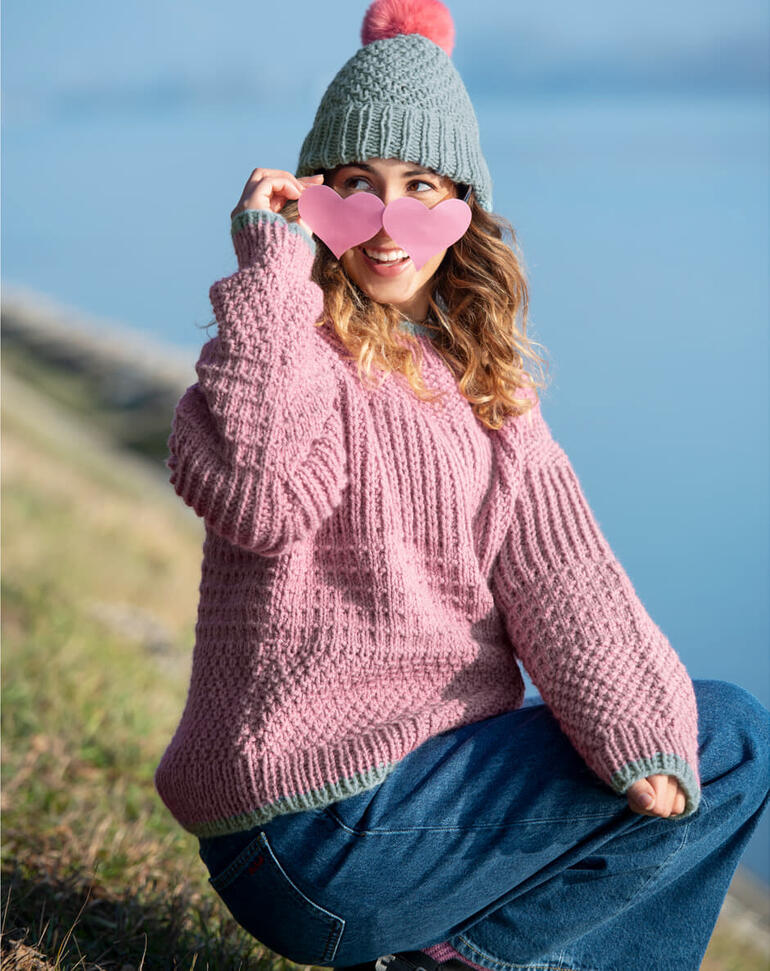 Frau in einem rosafarbenen Pullover und grau-grüner Mütze sitzt am Ufer.