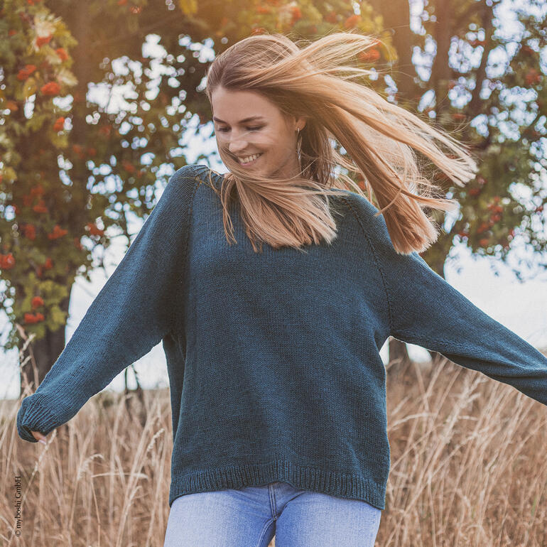 Frau in dunkelgrünem Pullover tanzt fröhlich auf einem Feld.