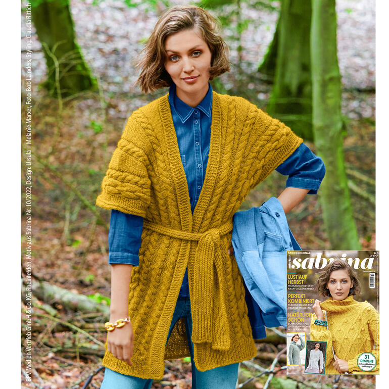 Frau trägt eine lange, gelbe Strickweste mit Gürtel und Zopfmuster, im Wald.