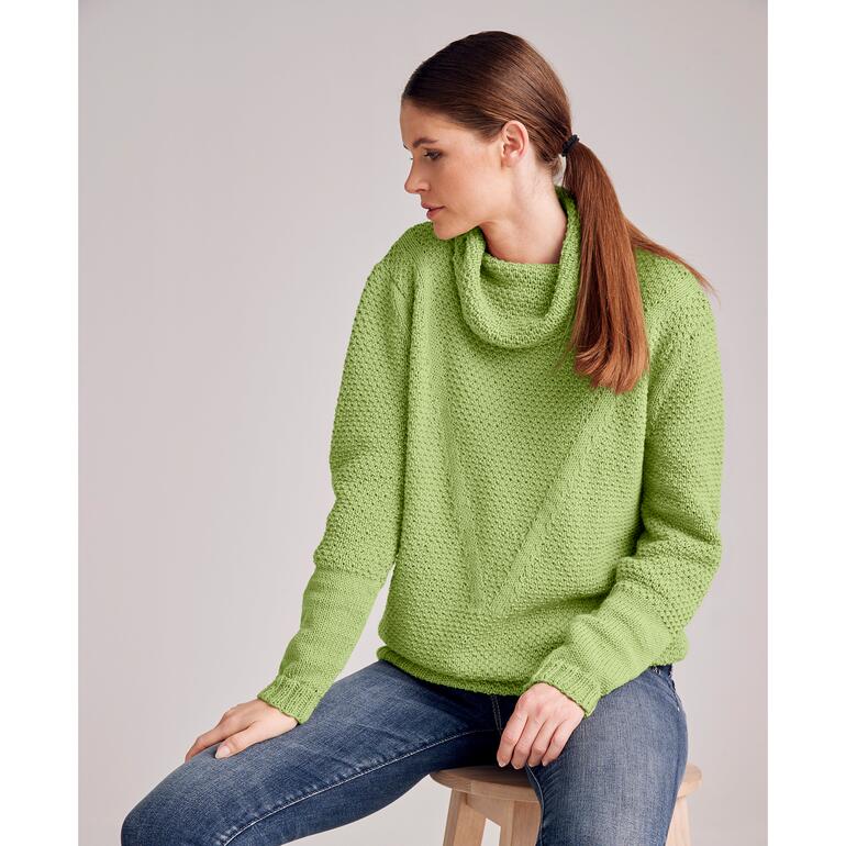 Grüner Pullover aus Merino-Cotton, Frau sitzt seitlich auf einem Hocker.