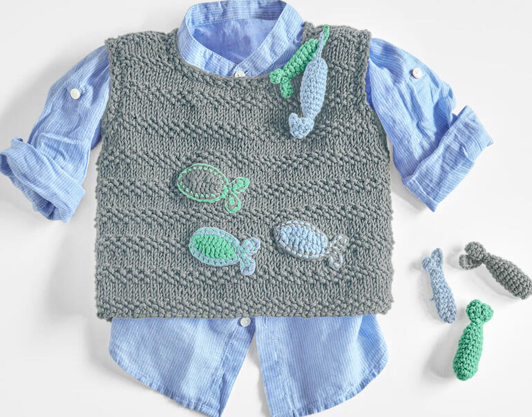 Grauer Baby-Pullunder mit gehäkelten Fischen, über einem blauen Hemd drapiert.