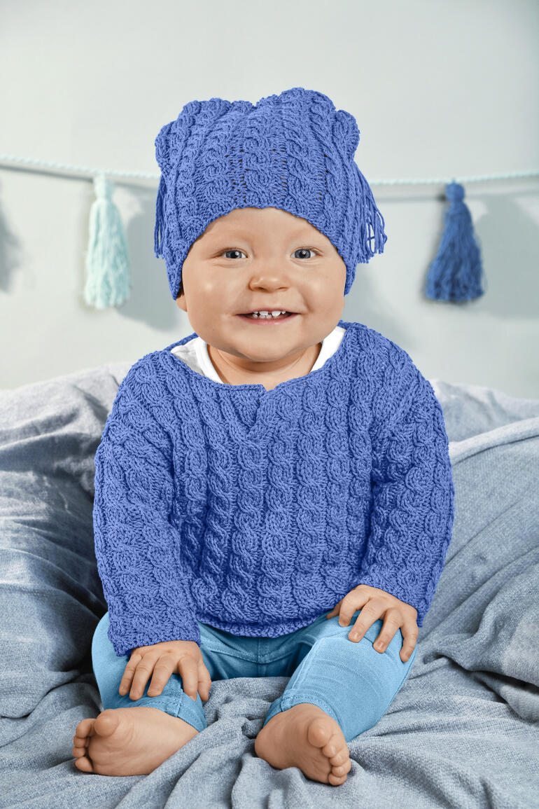 Lächelndes Baby in blauem Pulli mit Mütze auf einem hellen Hintergrund.