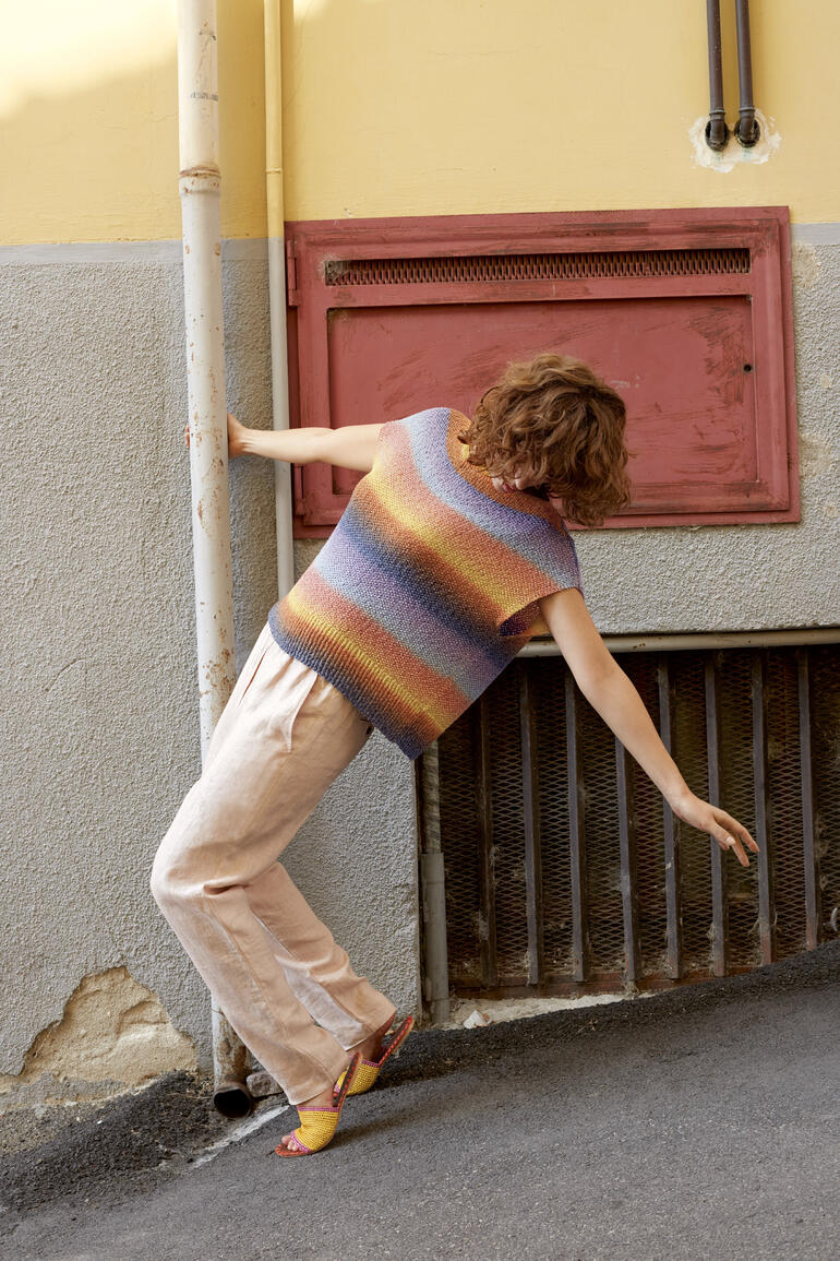 Frau trägt bunte, gestreifte Weste aus Paradise, lehnt an einer Wand.