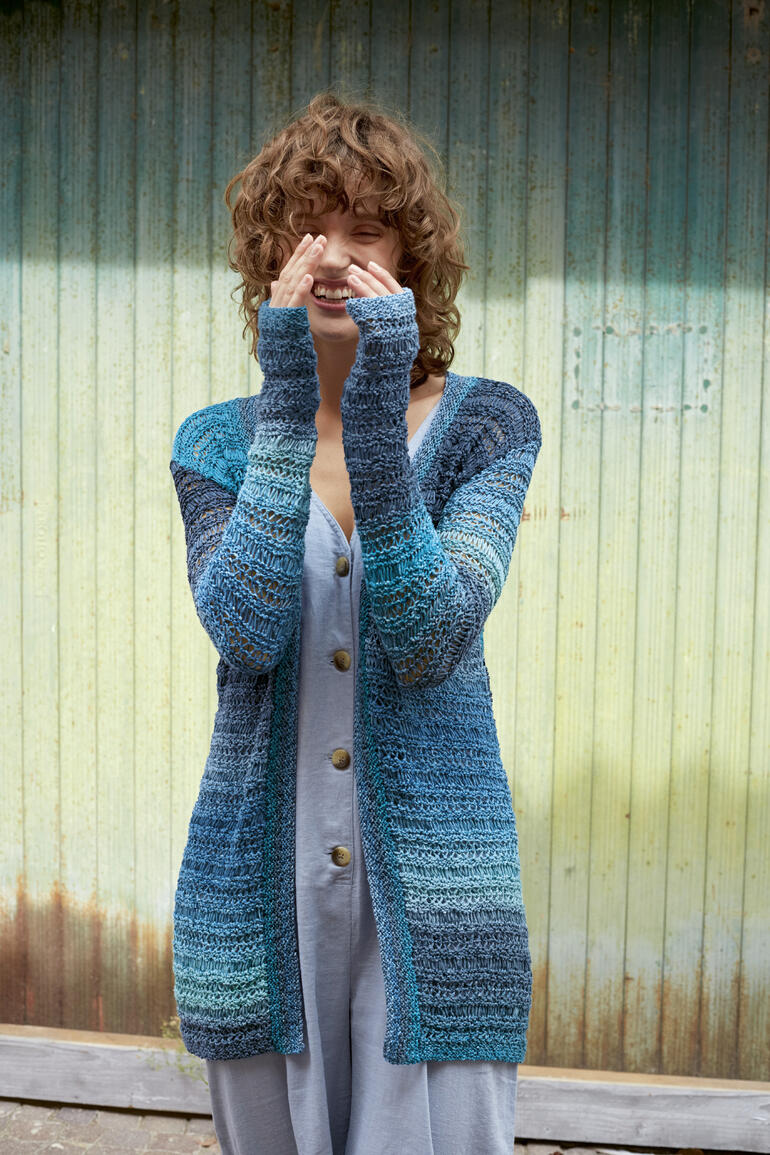Frau in blauer Long-Jacke aus Paradise, posiert vor einer farbenfrohen Wand.