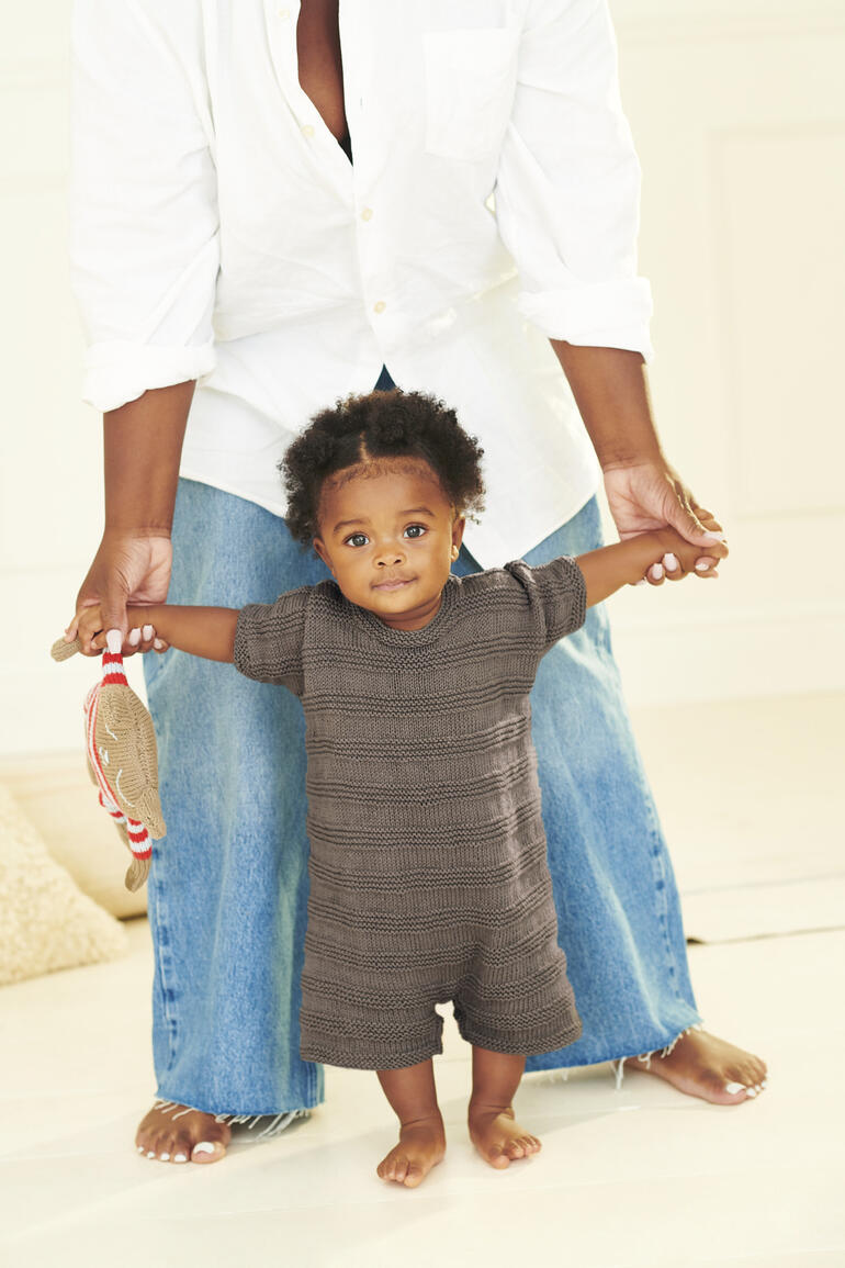 Babystrampler aus Bio-Baumwolle, getragen von einem kleinen Kind, das von einer Erwachsenen gestützt wird.