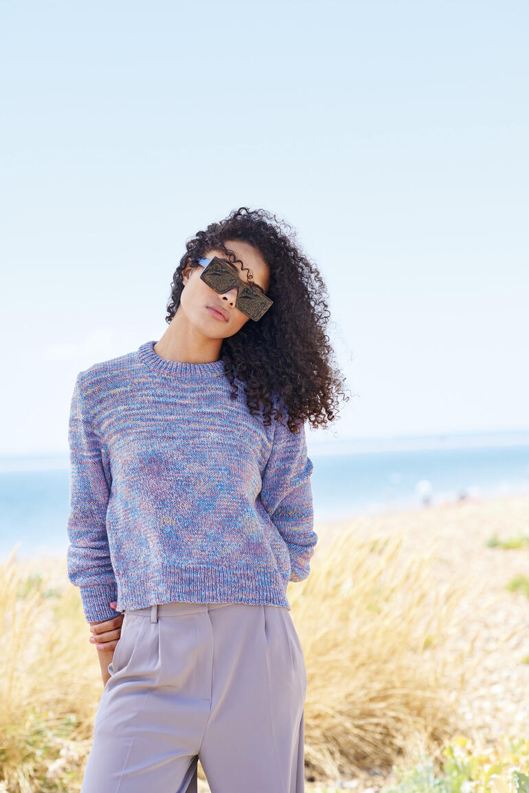 Pullover aus Creative Summer Sprinkles, getragen von einer Frau mit lockigem Haar und Sonnenbrille am Strand.