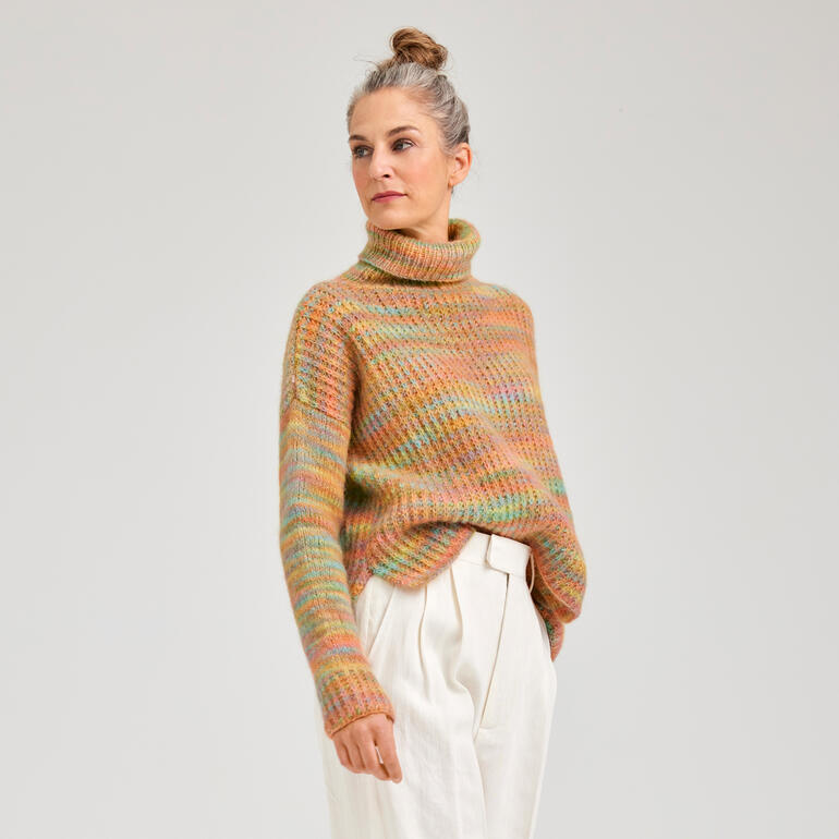 Frau in buntem Pullover aus Flou steht vor neutralem Hintergrund.